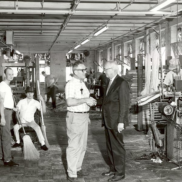 Black and white image of our former broom workshop in the 60's. Our foreman shaking hands with a gentleman in a suit in the center.